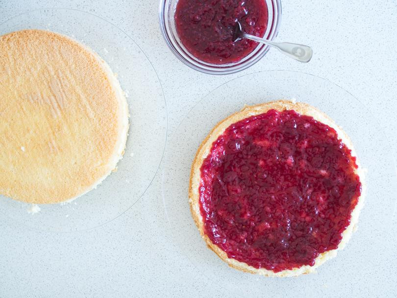 dwa blaty biszkoptu na szklanych talerzach, jeden z nich posmarowany konfiturą malinową, obok miseczka z konfiturą malinową i łyżeczką