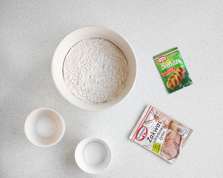 mąka pszenna, cukier i sól w miseczkach, opakowanie drożdży suszonych instant dr. oetkera oraz opakowanie zakwasu chlebowego żytniego dr. oetkera na blacie kuchennym