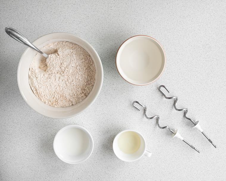 miska z mąką wymieszaną z drożdżami, zakwasem, solą i cukrem, obok miseczki z wodą, mlekiem i oliwą oraz mieszadła miksera