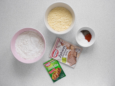 mąka pszenna chlebowa 750, mąka kukurydziana oraz przyprawy: sól, cukier, pieprz czarny i ostra papryka w miseczkach, obok opakowanie zakwasu żytniego dr. oetkera oraz opakowanie  drożdży suszonych instant dr. oetkera