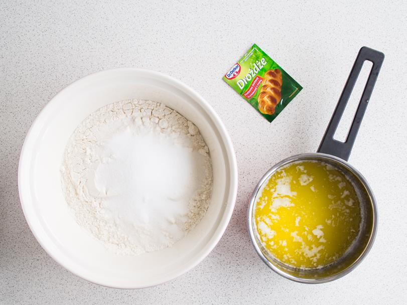 mąka i cukier w misce, obok opakowanie drożdży suszonych instant oraz rozpuszczone masło w rondelku