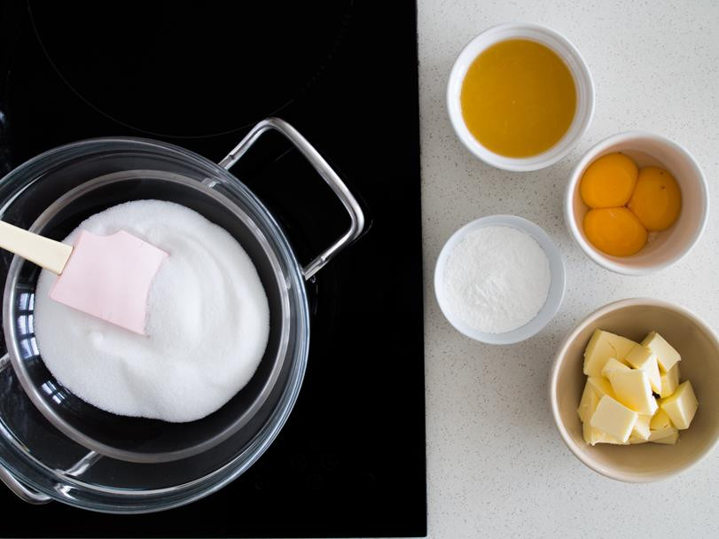 cukier w misce nad garnkiem z wodą na kuchence, obok sok z pomarańczy i cytryny, żółtka, skrobia kukurydziana oraz mało pokrojone w kostkę w osobnych miseczkach