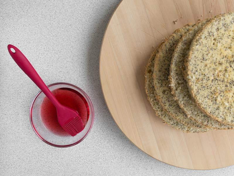 cztery blaty biszkoptu makowego na drewnianej desce powstałe do przekrojeniu dwóch biszkoptów na pół, obok szklanka z wódką żurawinową wymieszaną z wodą oraz pędzelek silikonowy do nasączenia biszkoptów