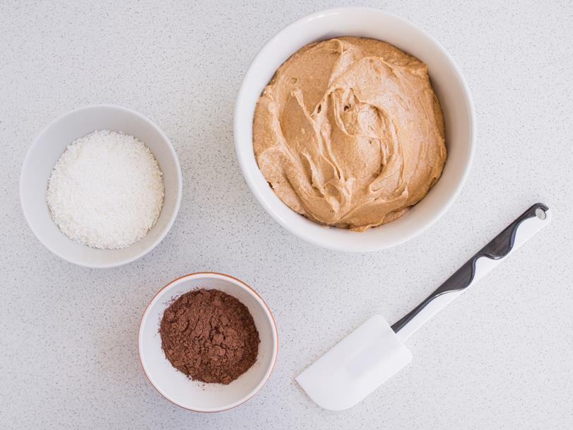 wiórki kokosowe, kakao i ciasto na babkę w osobnych miskach, obok szpatułka kuchenna do mieszania