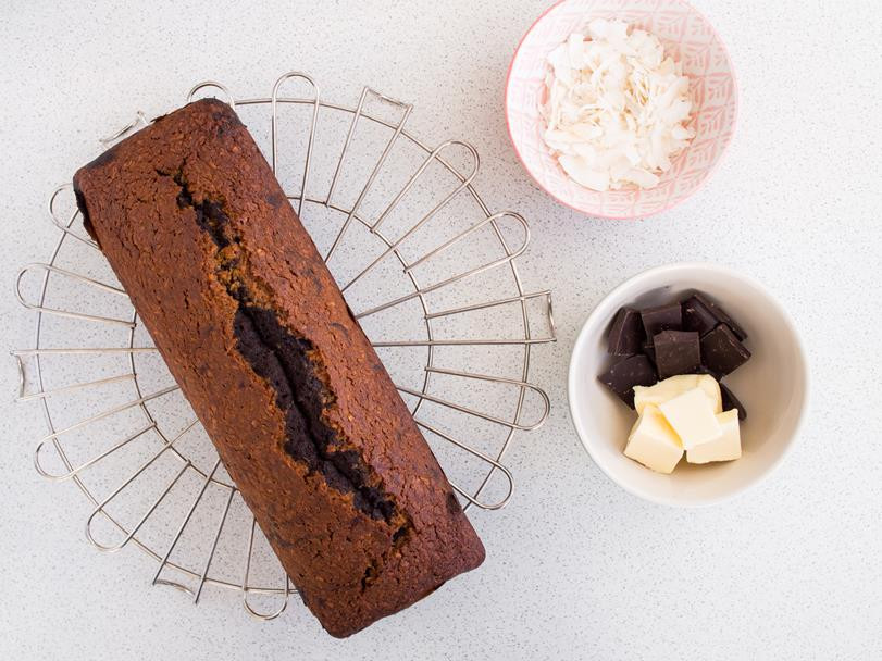upieczona babka kokosowo-czekoladowa na kratce do studzenia, obok miseczka z czekoladą gorzką i masłem oraz miseczka z płatkami kokosa