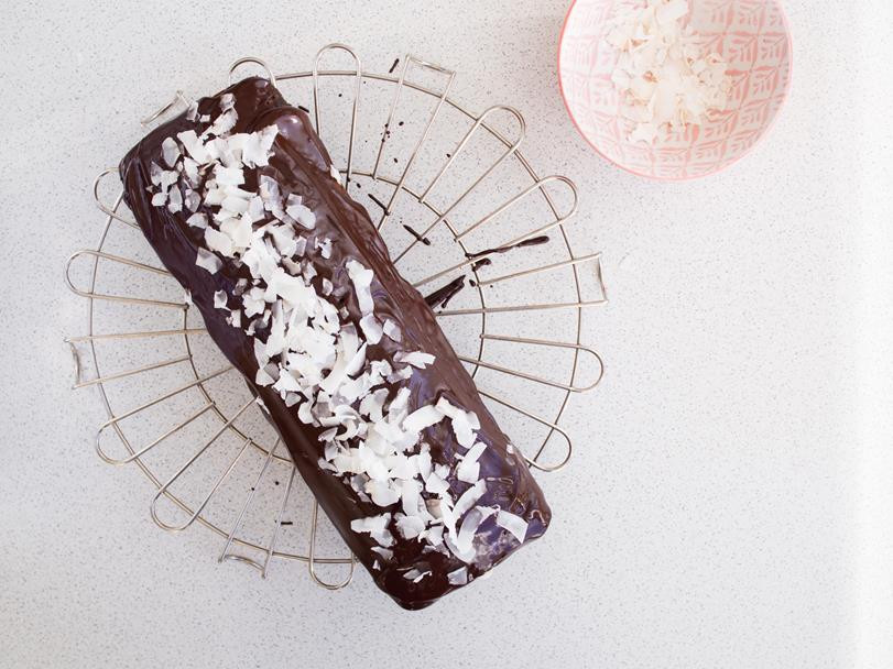 upieczona babka kokosowo-czekoladowa na kratce do studzenia, polana czekoladą i posypana płatkami kokosa, obok miseczka z płatkami kokosa