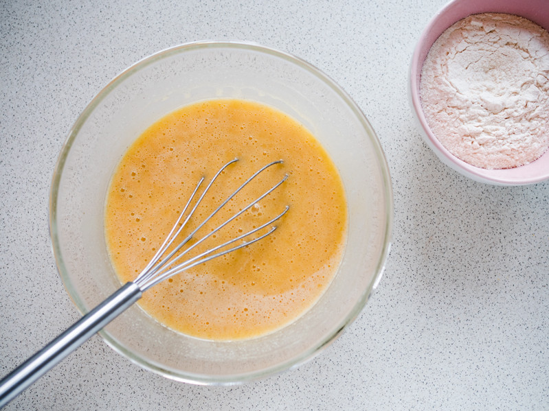 masa jajeczna w szklanej misce z rózgą kuchenną i mąka z proszkiem do pieczenia w drugiej miseczce