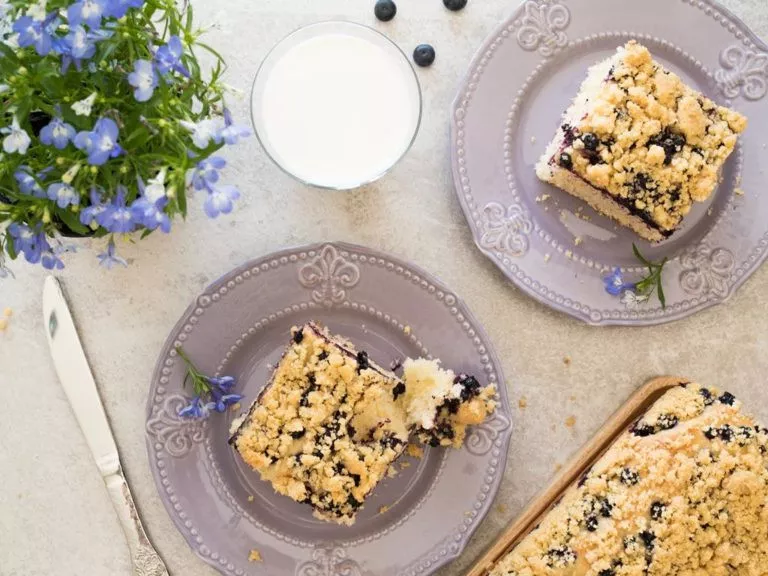 Ciasto drożdżowe z jagodami podane na fioletowych talerzykach, Obok w szklance mleko i kwiaty.