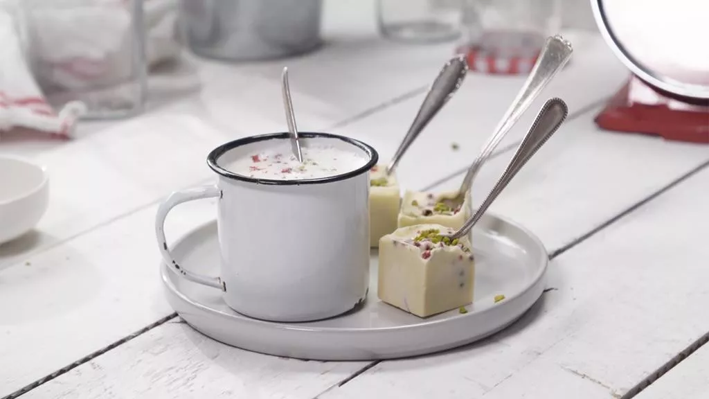 biała czekolada z różowym pieprzem i pokruszonymi pistacjami zatknięta na łyżeczce, obok garnuszek z gorącym mlekiem i rozpuszczoną w nim czekoladą - Wszystkiego Słodkiego