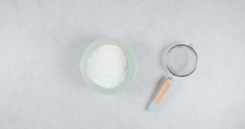 mąka pszenna, ziemniaczana i proszek do pieczenia w misce, obok sitko do ich przesiania