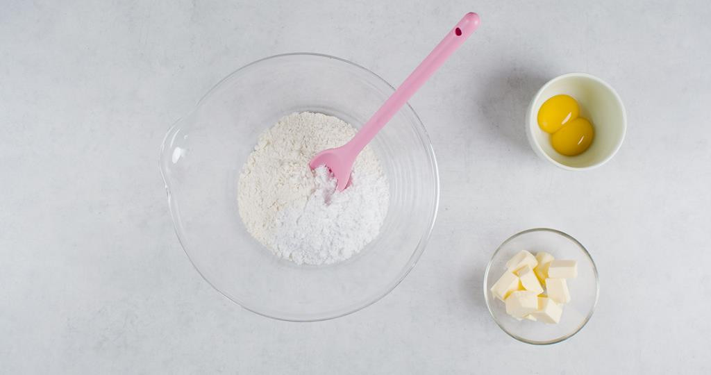 Mąka z cukrem pudrem w szklanej misce, obok masło pokrojone w kostkę i żółtka w osobnych miseczkach