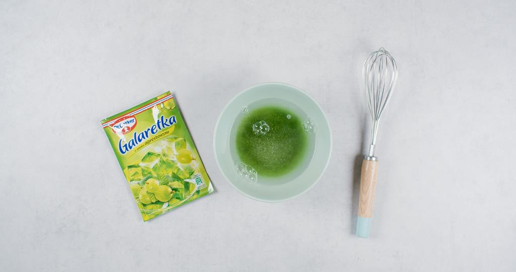 opakowanie galaretki agrestowej dr. oetkera, obok miseczka z wodą i wsypaną do niej galaretką agrestową oraz ubijaczka kuchenna (rózga)