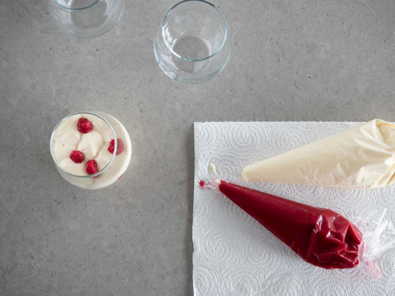 krem malinowy i śmietanowy w rękawach cukierniczych na ręczniku papierowym obok deser z białej czekolady z malinami