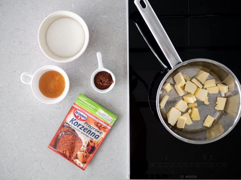 Pokrojone masło w garnku. Obok przyprawa korzenna, miód, cukier oraz kakao.