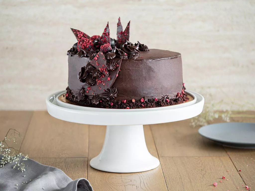 czekoladowy tort wegański z ozdobami z czekolady i liofilizowanymi malinami na bialej, ceramicznej paterze - Wszystkiego Słodkiego