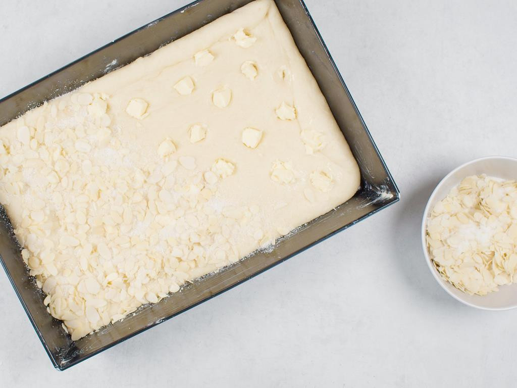 forma z ciastem drożdżowym posypanym mieszanką cukru i płatków migdałowych, obok miseczka z płatkami migdałowymi z cukrem