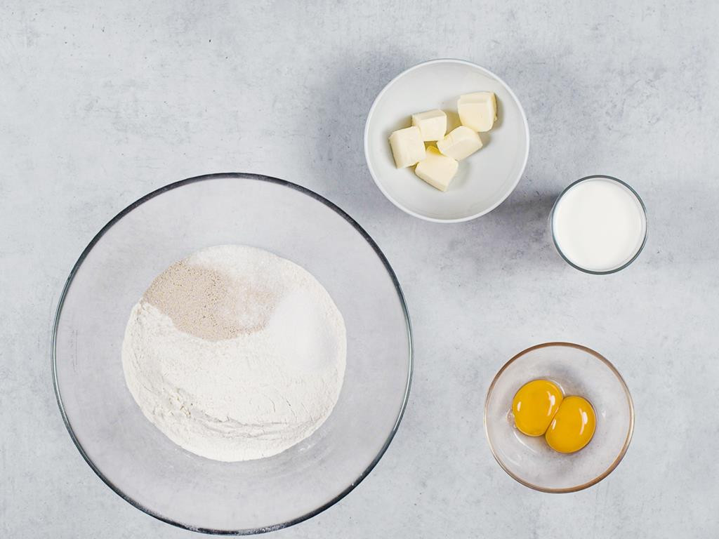 Mąka, cukier i suszone drożdże w misce, masło pokrojone w kostkę, żółtka i mleko w osobnych miseczkach