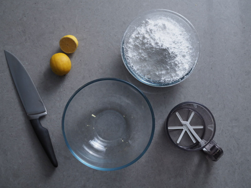 w miseczce przesiany cukier puder, szklana miska, przesiewacz do cukru, przekrojona cytryna oraz noż