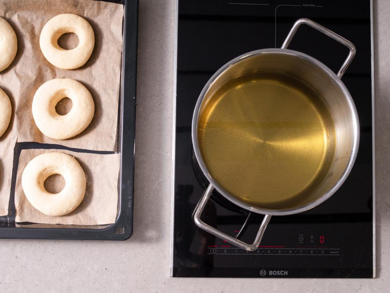 Wyrośnięte oponki na papierze do pieczenia. Obok olej rzepakowy w garnku.
