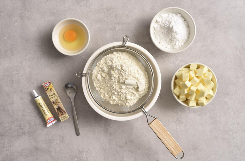 Mąka pszenna w misie. Obok pokrojone masło, cukier puder, pasta z wanilii Dr. Oetkera oraz jajko.