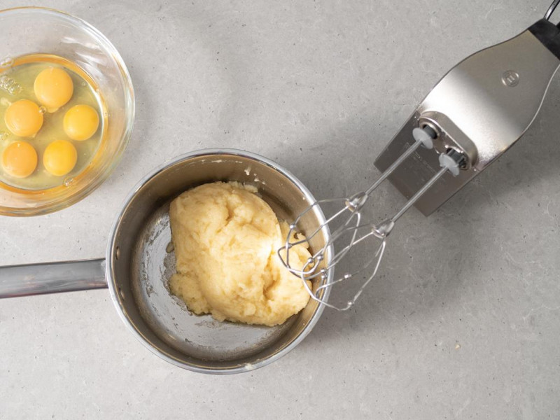 W misce zaparzone ciasto ptysiowe, obok mikser ręczny firmy Bosch i jajka w miseczce.