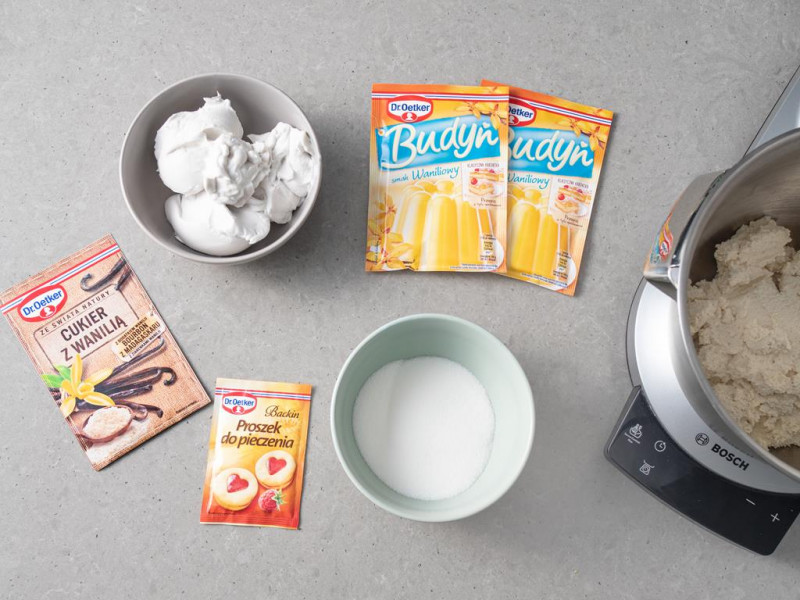 Na blacie budyń o smaku waniliowym Dr. Oetkera, proszek do pieczenia, cukier z wanilią, cukier w miseczce i mleko kokosowe.
