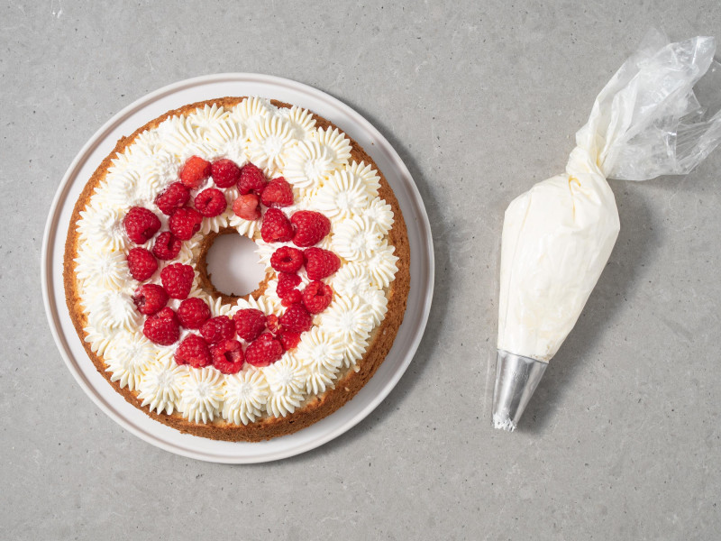 maliny ułożone na środku kremu waniliowego na cieście anielskim, obok rękaw cukierniczy z kremem