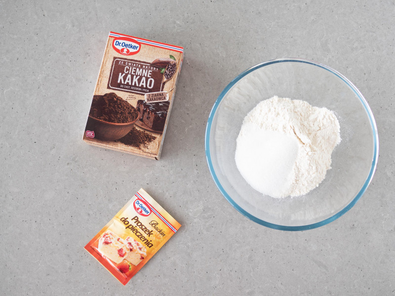 W szklanej misie mąka pszenna, cukier i sól. Obok proszek do pieczenia Dr. Oetkera i Ciemne Kakao Dr. Oetkera.