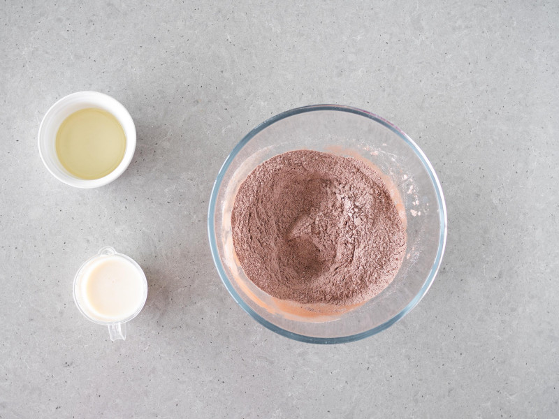 W szklanej misie wymieszane suche składniki na czekoladowe ciasto wegańskie. Obok napój sojowy i olej rzepakowy.