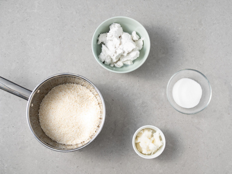 W garnku wiórki kokosowe, obok olej kokosowy, mleczko kokosowe i cukier.