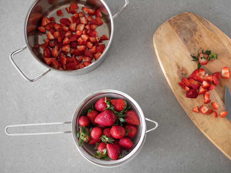 Umyte truskawki na sitku i obok pokrojone owoce w garnku.
