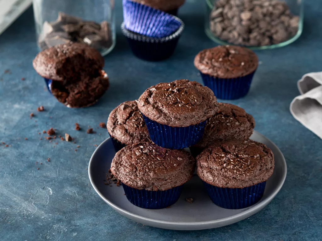 Czekoladowe muffinki z kawałkami czekolady w metalicznych papilotkach na szarym talerzyku.