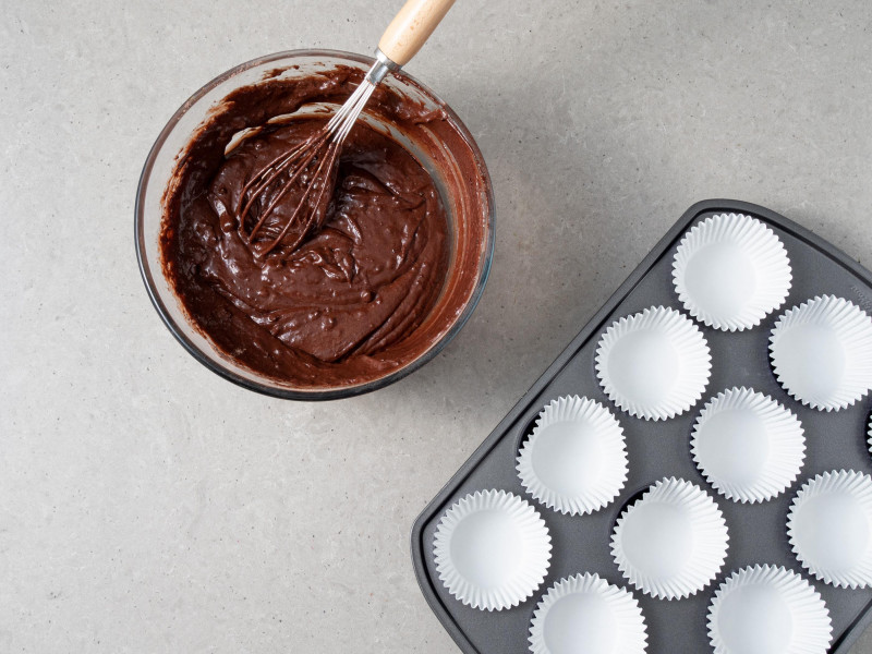 W misce szklanej ciasto czekoladowe na muffinki. Obok foremka na muffinki wype艂niona papilotkami.