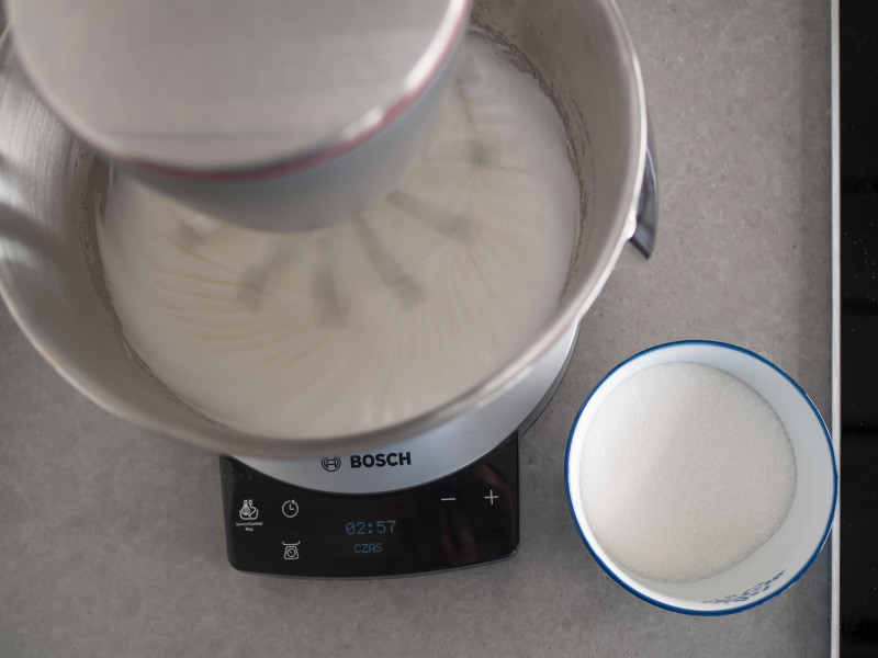 białka ubijane w robocie kuchennym marki bosch na sztywną pianę, obok miseczka z cukrem