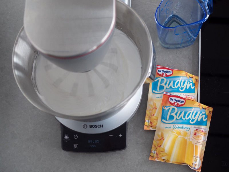 białka ubite z cukrem w robocie kuchennym marki bosch, obok dwa opakowania budyniu waniliowego od dr. oetkera oraz olej w niebieskim dzbanku z miarką kuchenną