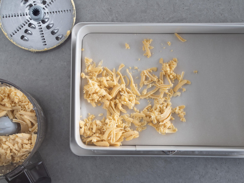 prostokątna forma do pieczenia wyłożona papierem do pieczenia z niewielką ilością startego ciasta kruchego, reszta startego ciasta obok w malakserze