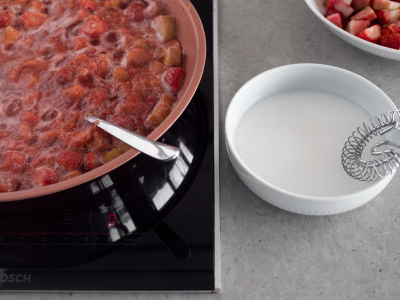 miękkie truskawki z rabarbarem na patelni, obok miseczka z mąką ziemniaczaną rozmieszaną z wodą