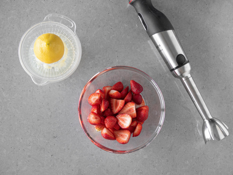 W miseczce pokrojone truskawki, obok cytryna i blender ręczny firmy Bosch.
