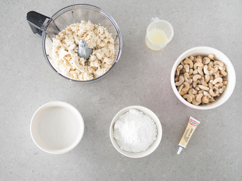 W pojemniku malaksera pokruszony serek tofu. Obok namoczone orzechy nerkowca, cukier puder, pasta z wanilii Dr. Oetkera, sok z cytryny i mleczko kokosowe.