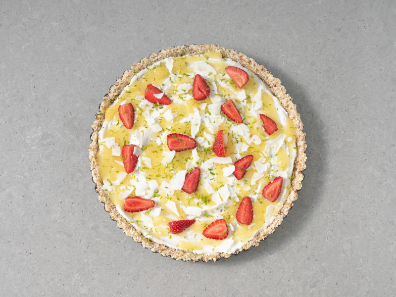 Tarta bez pieczenia pina colada polana dżemem, posypana płatkami kokosa i świeżymi truskawkami.