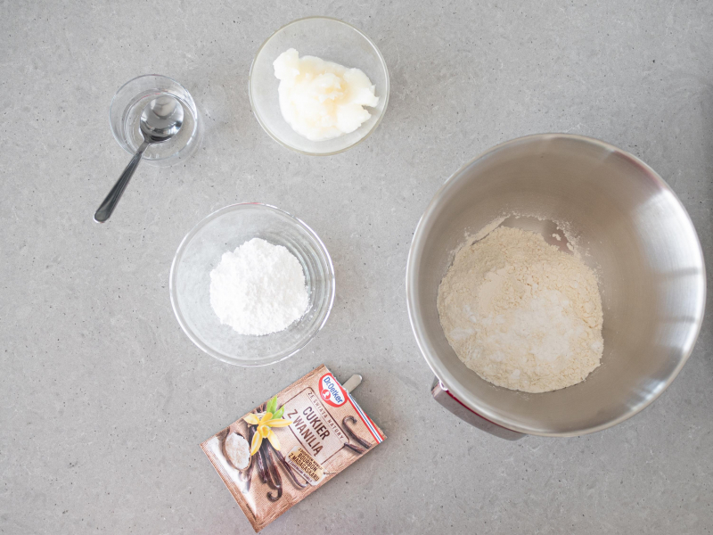 Mąka pszenna w misie. Obok w miseczkach mąka ziemniaczana, cukier puder, olej kokosowy i zimna woda. W opakowaniu cukier z wanilią Dr. Oetkera.
