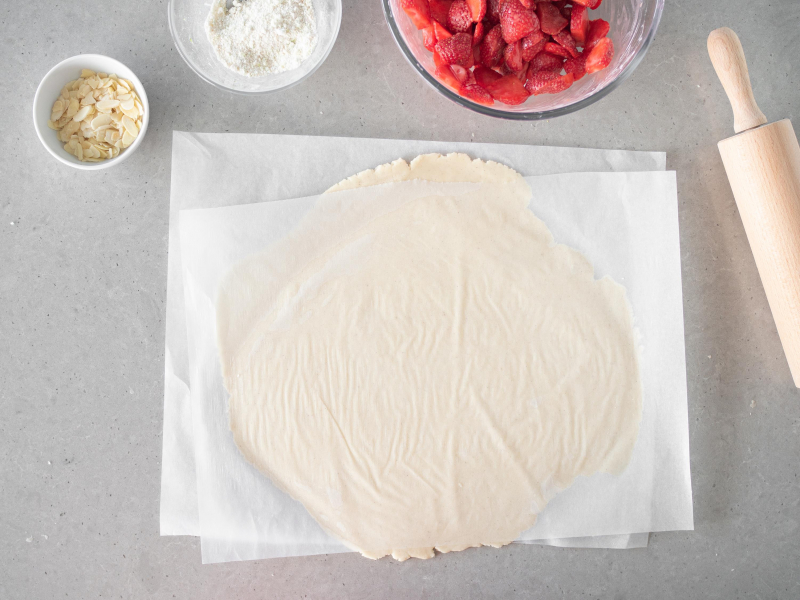 Rozwałkowane pomiędzy arkuszami papieru kruche ciasto wegańskie. Obok w misce wałek, płatki migdałów i truskawki wymieszane z mąką ziemniaczaną.