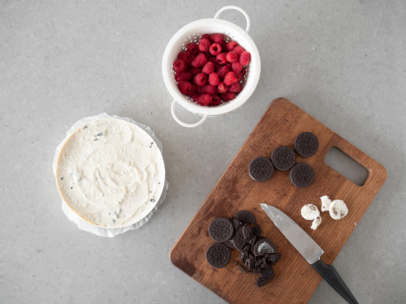 Na drewnianej desce ciasteczka oreo. Obok maliny i masa serowa w tortownicy.