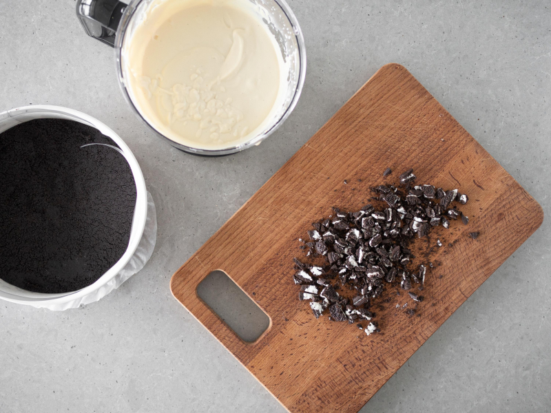 Na drewnianej desce pokruszone ciasteczka oreo. Obok przygotowany wcześniej spód i masa a'la serowa w malakserze.