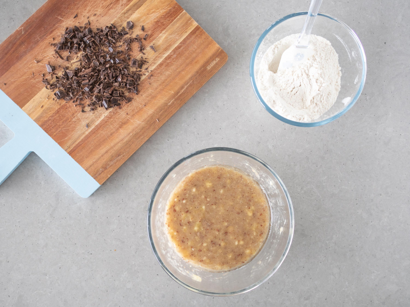 Pokrojona gorzka czekolada na drewnianej desce. Obok wymieszane mokre i suche składniki.