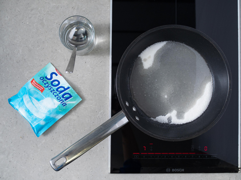 Cukier z wodą na patelni. Obok soda oczyszczona Dr. Oetkera i woda w szklance.