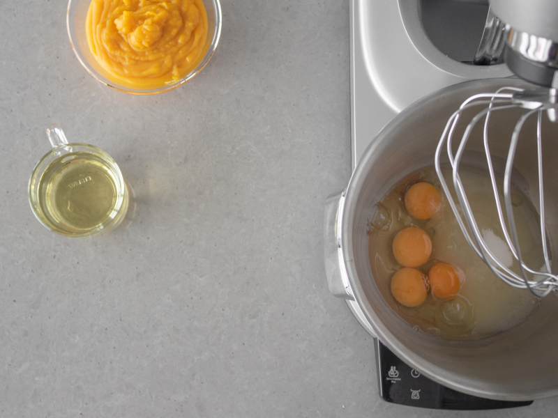 W misie robota kuchennego firmy Bosch jajka z cukrem i ekstraktem pomarańczowym. Obok puree z dyni w misce i olej w szklance.