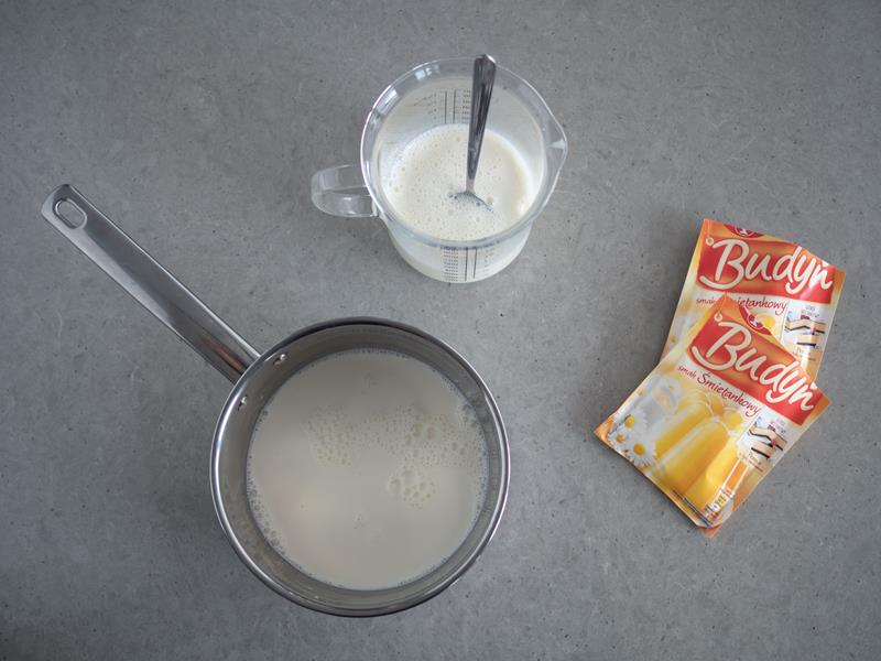 w rondelku mleko do zagotowania na budyń, obok dwa opakowania budyniu śmietankowego dr. oetkera oraz mleko w miarce kuchennej z łyżką do rozmieszania z budyniami