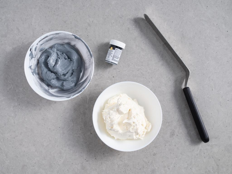 dwie miseczki z kremem maślanym - jedna z dodatkiem białego barwnika, druga z dodatkiem czarnego barwnika, dzięki któremu krem ma szarą barwę, obok szpatuła do rozprowadzania kremu oraz opakowanie czarnego barwnika spożywczego Wilton