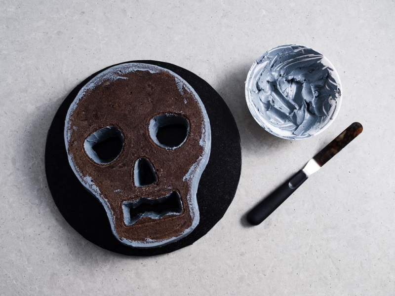 miseczka z szarym kremem maślanym, szpatuła do kremu oraz ciasto czekoladowe w kształcie czaszki z bokami posmarowanymi szarym kremem maślanym